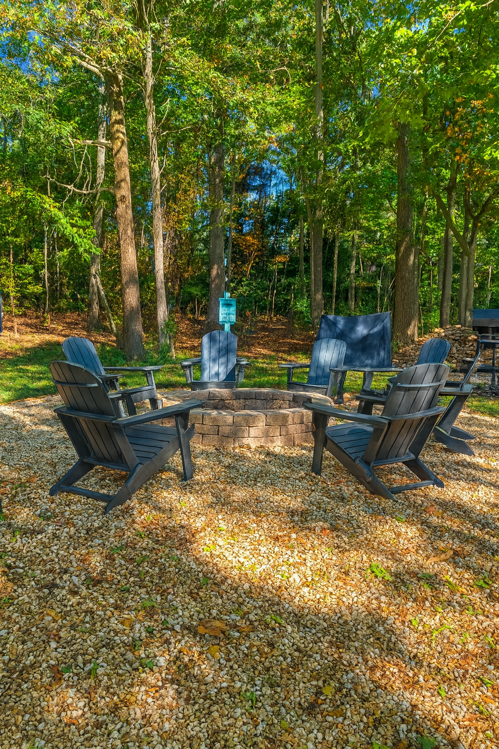 Fire pit with Adirondack chairs in a wooded area with gravel ground.