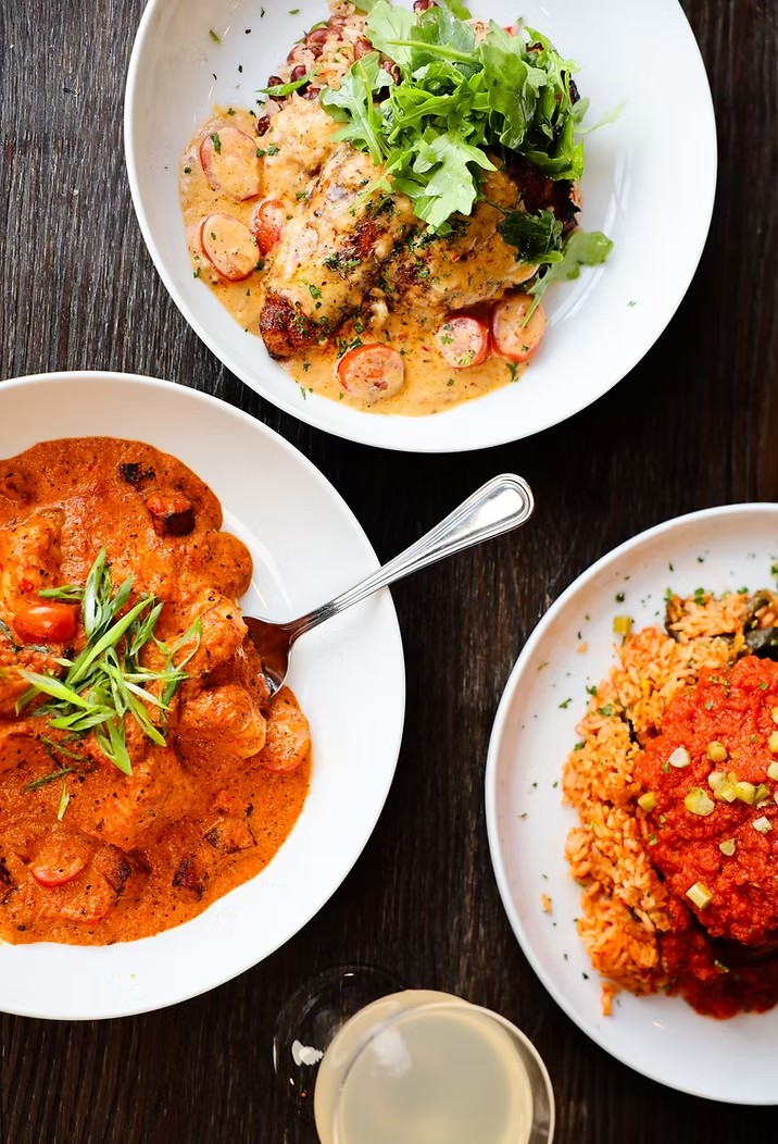 Three plates of food on a dark wooden table, including a creamy dish, tomato-based stew, and rice.