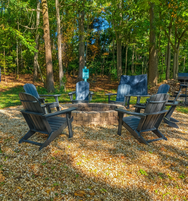 Fire pit with six blue chairs in a wooded area, surrounded by gravel.