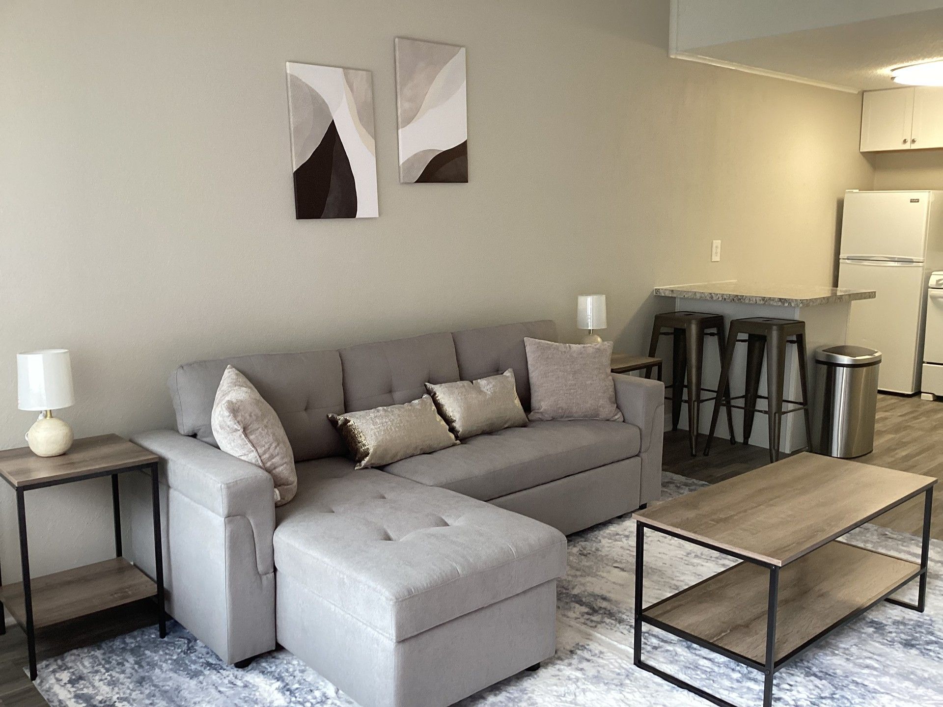 Living room with gray sectional, coffee table, and bar stools. Artwork on wall, and small lamps.