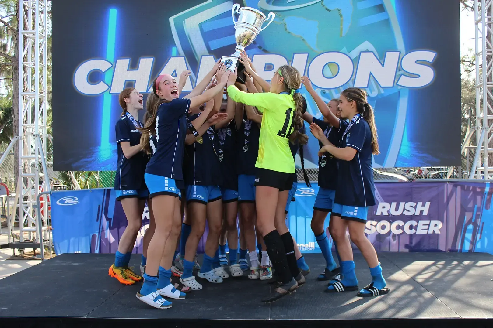 Soccer team celebrating championship victory, holding trophy high. Bright stage,