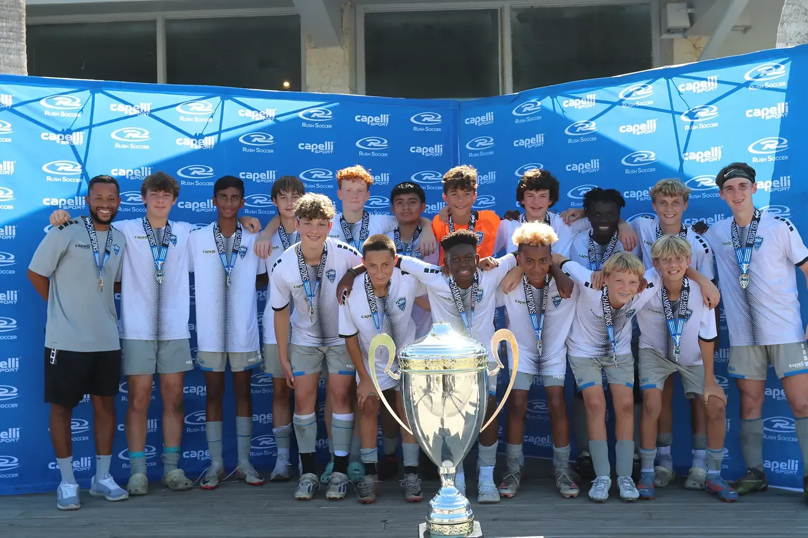 Soccer team posing with trophy, wearing medals, in front of a blue backdrop, celebrating victory.