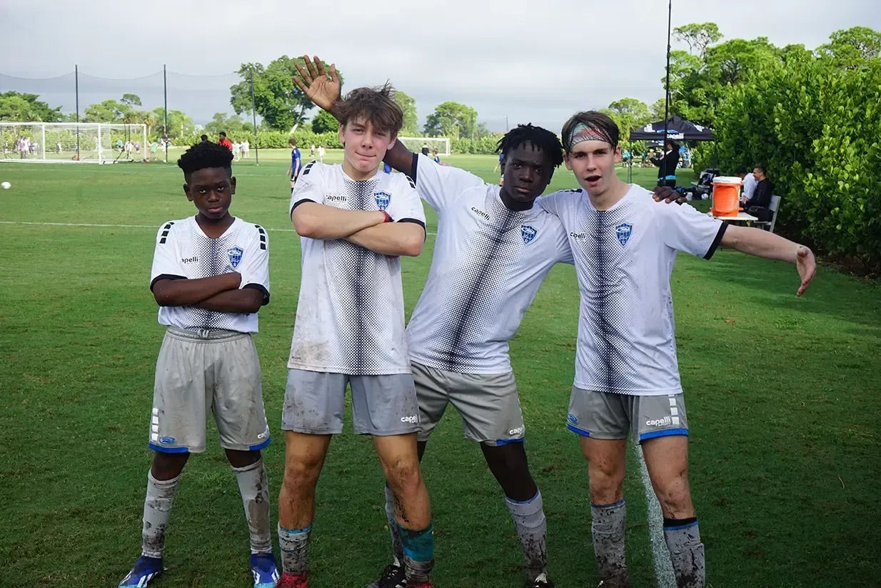Soccer players pose for a photo on a field, covered in mud. Three smile, one raises arm.
