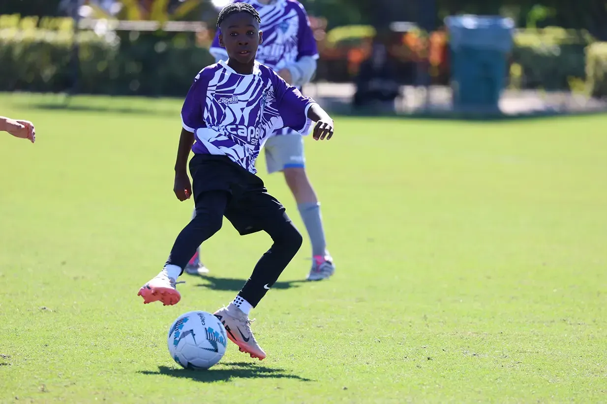 Boy in purple soccer jersey dribbles a soccer ball on a green field.