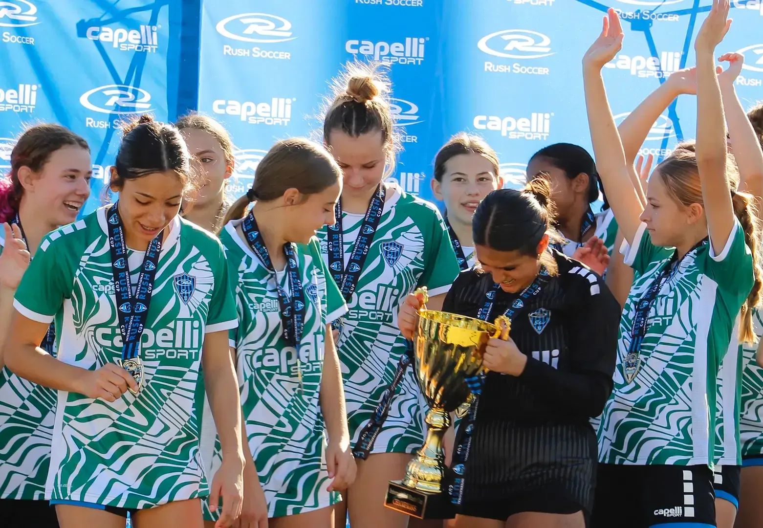 Soccer team celebrating victory with a trophy. Players wear green and white jerseys, some raising arms in excitement.