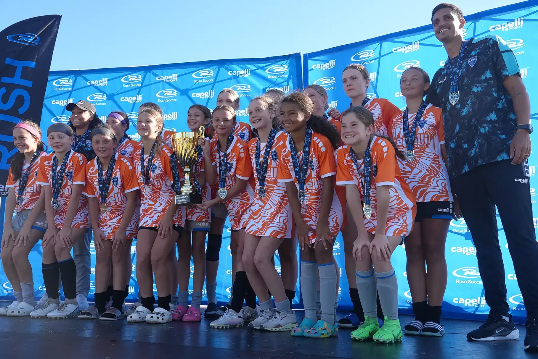 Soccer team in orange and white jerseys with medals, holding a trophy, posing on a stage.