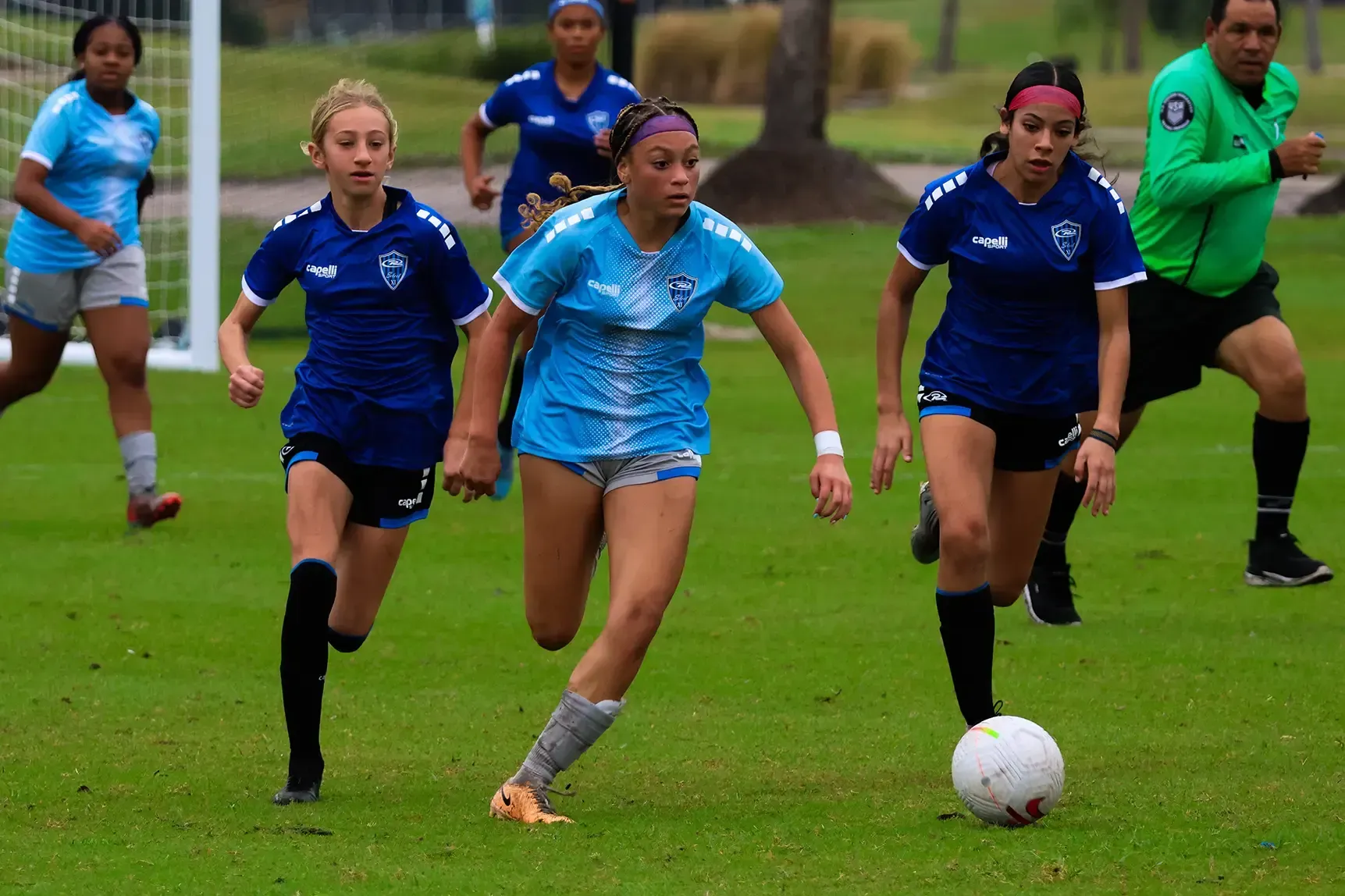 Soccer players in blue and light blue jerseys running on a green field, ball in play.