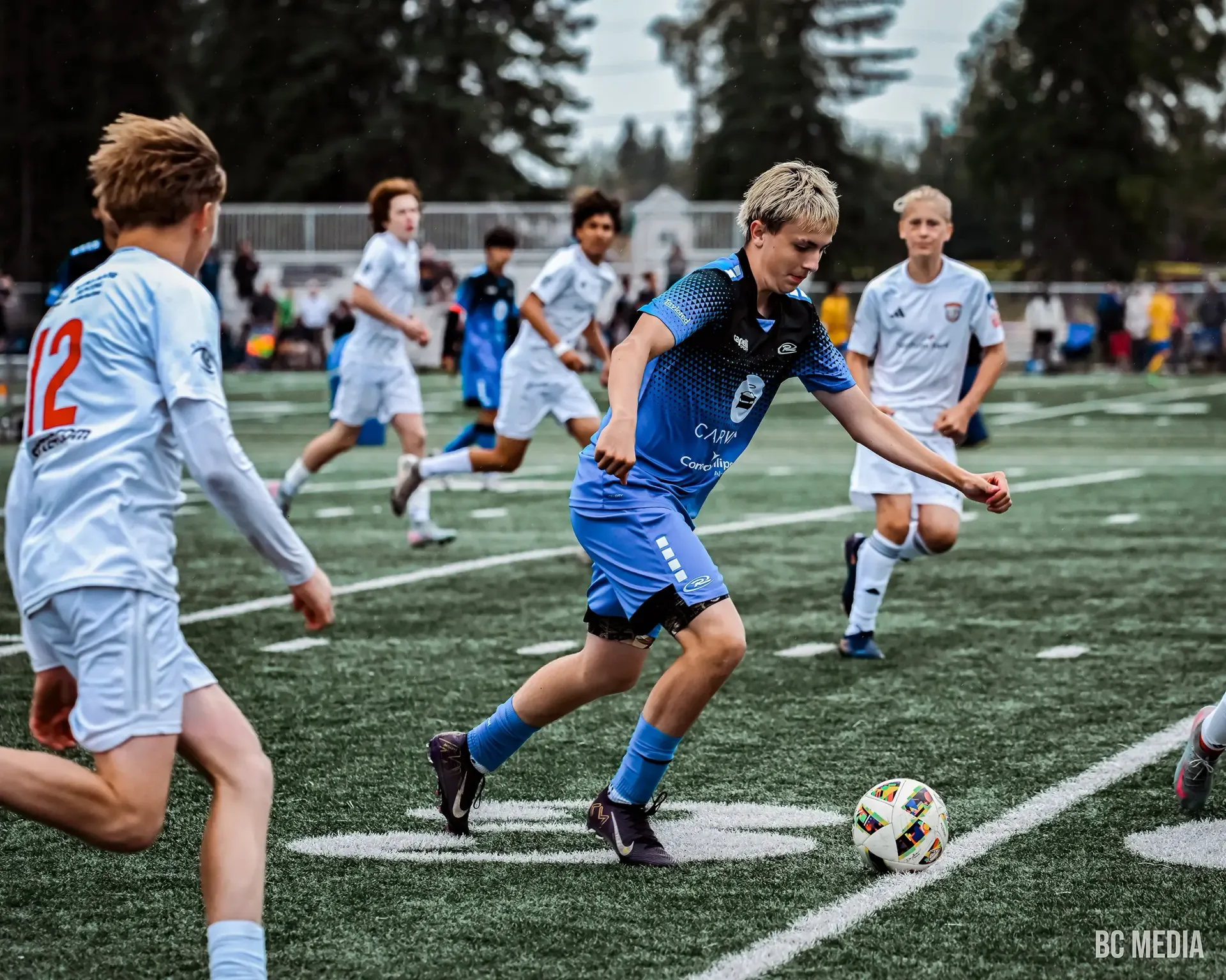 Soccer players in blue and white jerseys on a green field. One player dribbles the ball toward the goal.