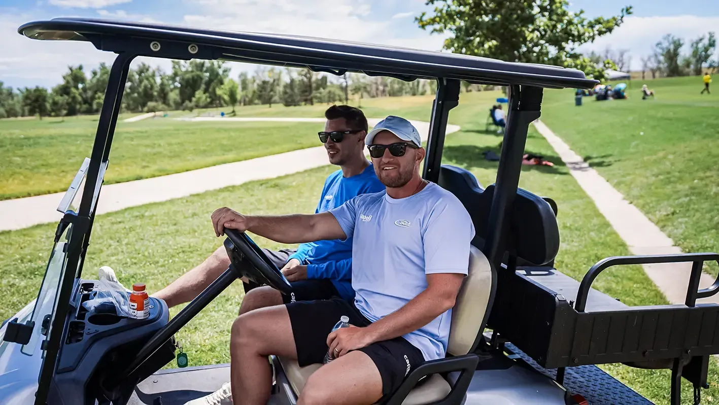 Two men in sunglasses driving a golf cart on a green grassy field, sunny day.