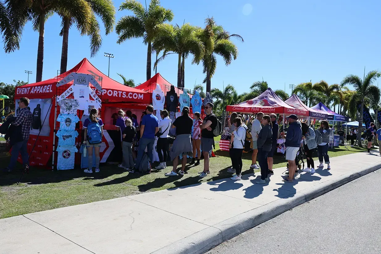 Crowd gathered at outdoor booths, selling merchandise under red tents on a sunny day.
