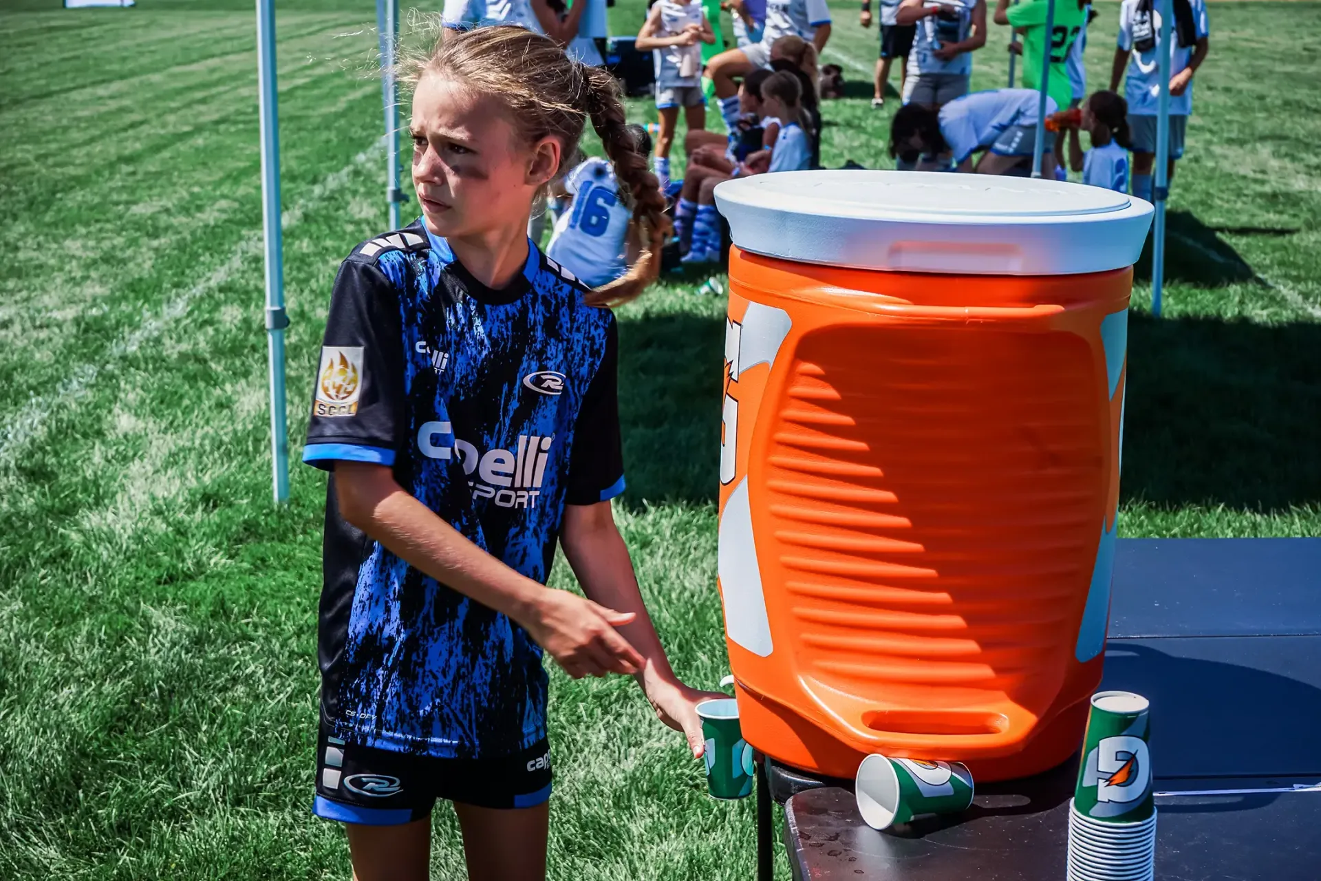 Girl in blue jersey at a water cooler, pointing to a cup. Outdoors, sunny, with other people.