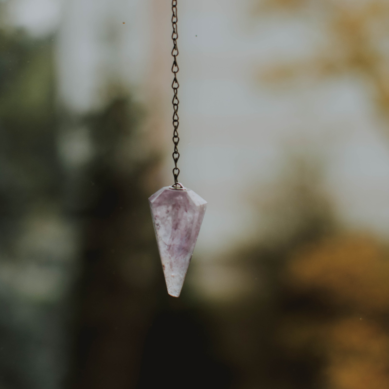 A pointed, translucent light purple crystal pendulum hangs from a delicate metal chain against a soft, blurry background.