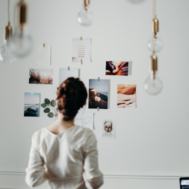 A person with their back to the camera looks at photos and notes pinned to a white wall in a room with hanging bulbs.