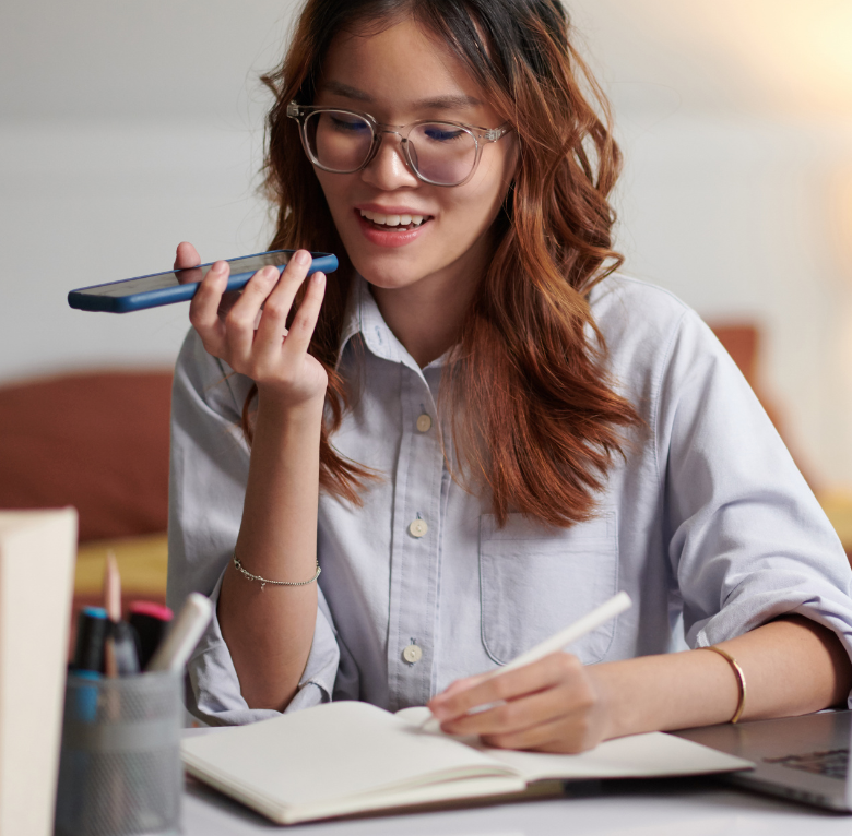 A person wearing glasses uses a smartphone to record a voice message while writing in an open notebook at a desk.