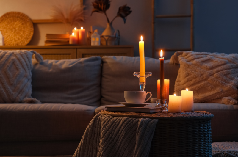 A cozy living room setting with glowing candles on a small round table, soft lighting, and pillows on a sofa.