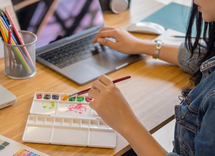 A person works at a desk, using a paintbrush on a watercolor palette while typing on a laptop with their other hand.