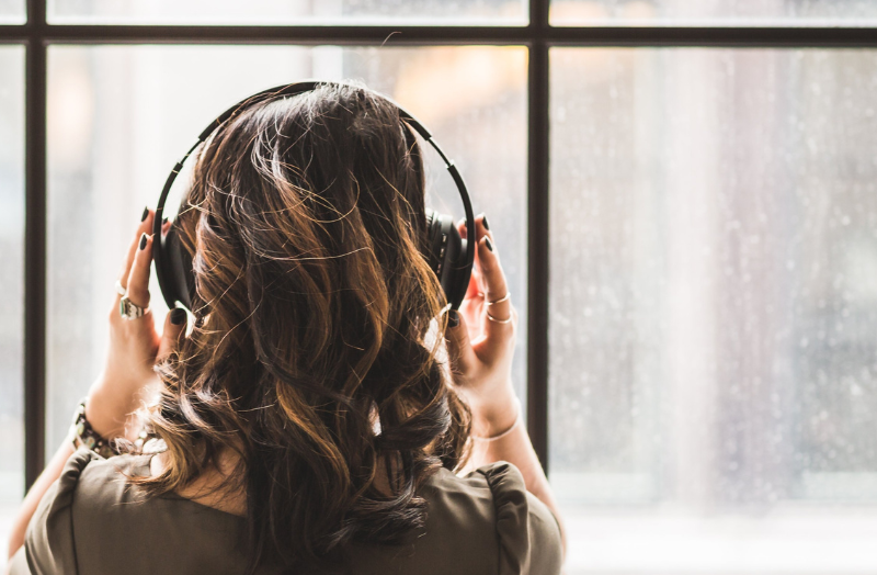 A person with wavy brown hair seen from behind, wearing headphones and looking out a window.