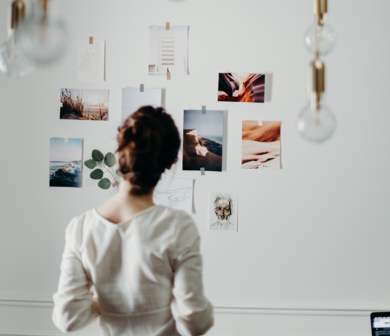 A person stands before a white wall covered with various photos and art prints, appearing to study a mood board.