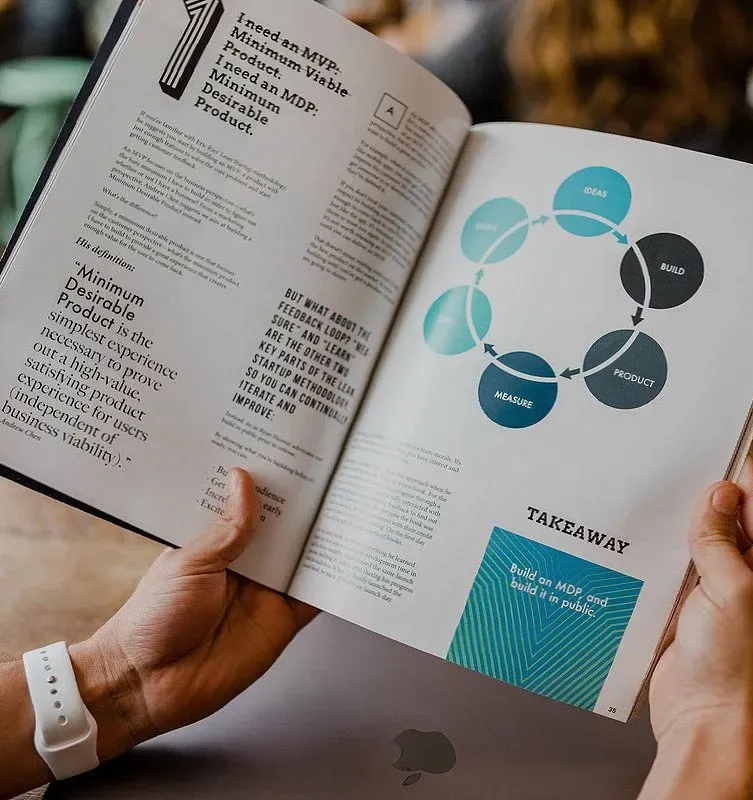 Hands hold an open book showing a business lifecycle diagram with blue and dark gray circular icons on a wooden surface.
