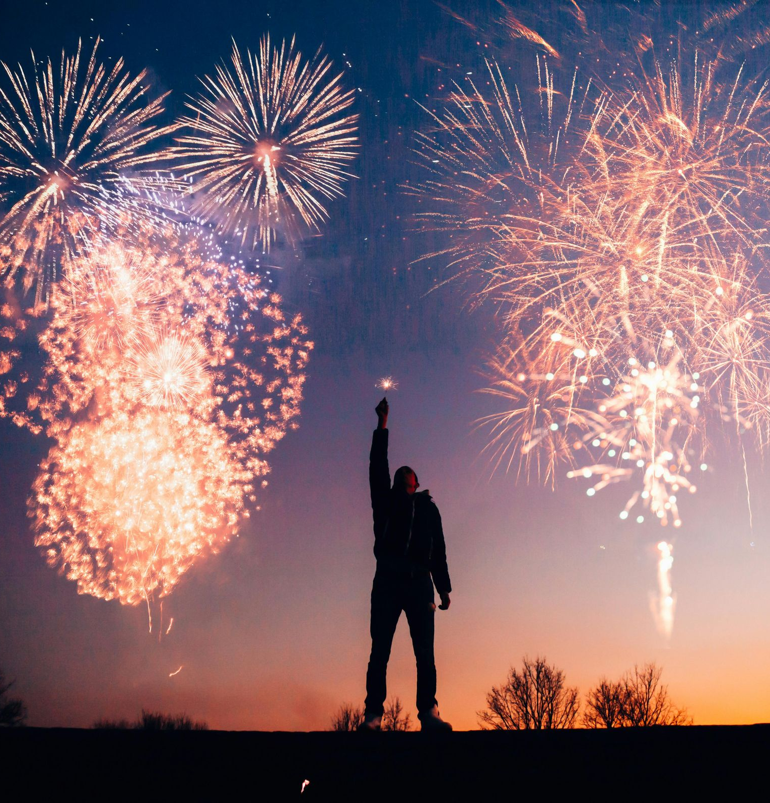 A silhouette of a person standing on a hill at sunset, holding their arm up toward bright fireworks in the night sky.