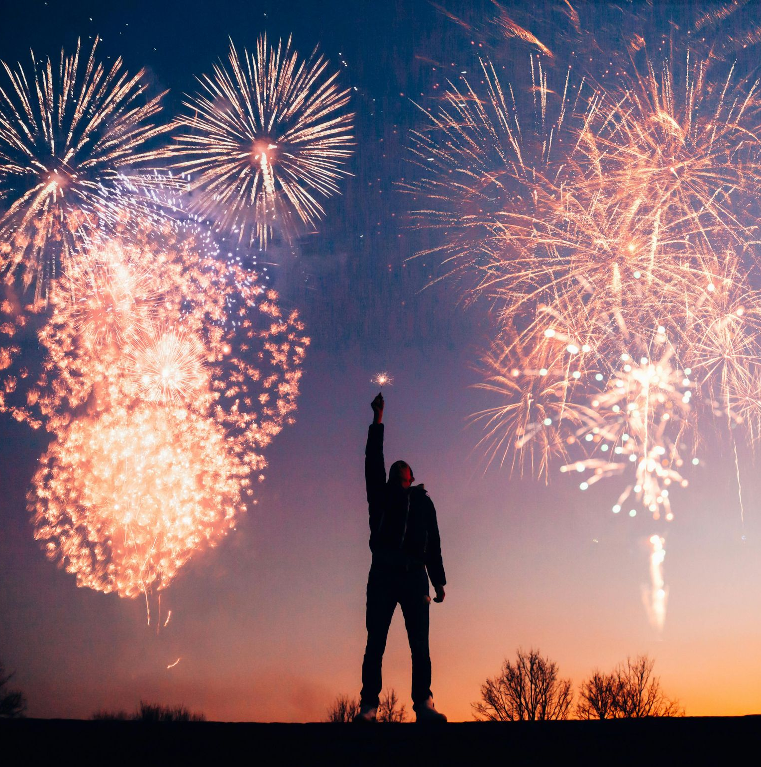 A silhouette stands with an arm raised towards a night sky filled with bright, multi-colored fireworks.