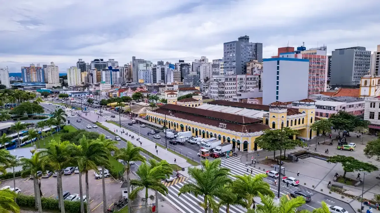 Vista aérea de uma cidade com prédios altos, um mercado, palmeiras e um céu nublado.