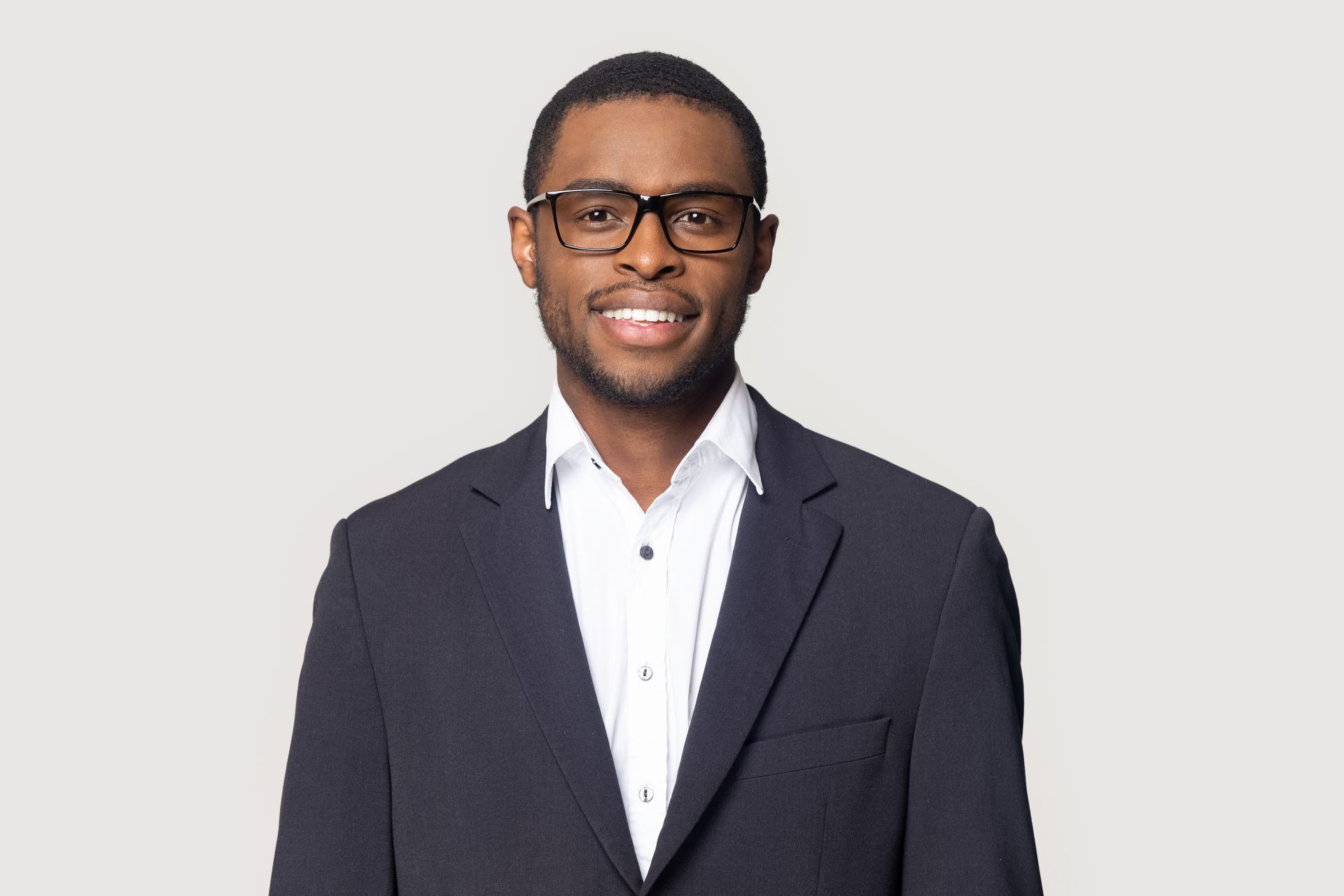 Man in a suit and glasses smiles at the camera. He is in front of a neutral backdrop.