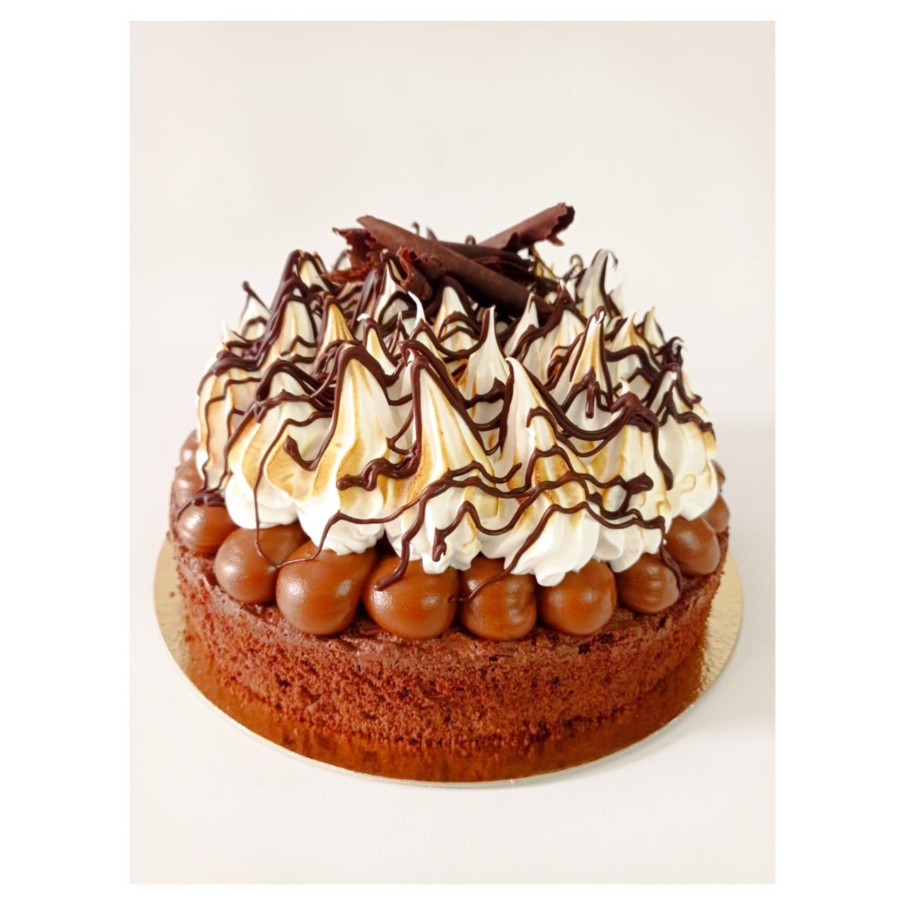 Pastel de chocolate cubierto con caramelo, merengue y virutas de chocolate, sobre una base de pastel dorada.