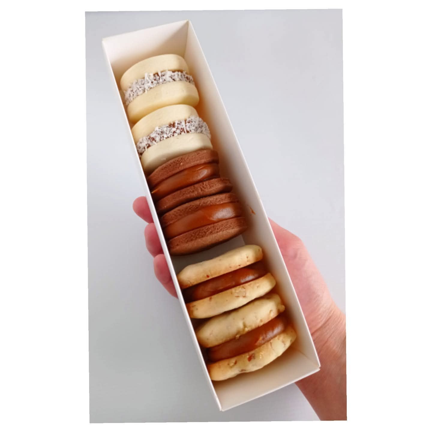 Mano sosteniendo una caja blanca llena de seis alfajores, algunos con glaseado blanco y coco, otros con relleno de caramelo.