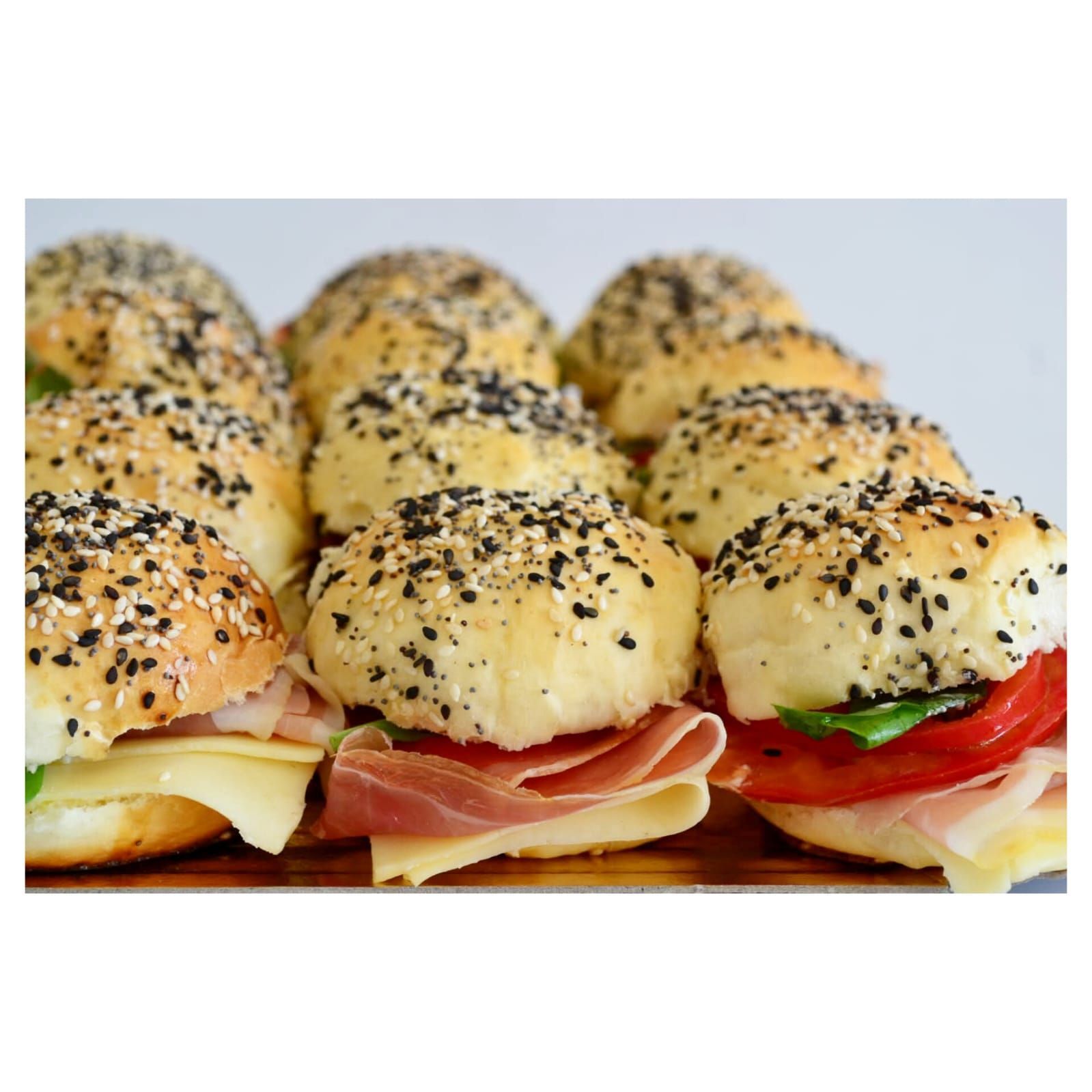 Mini sándwiches en pan con semillas, con jamón, queso, tomate y albahaca sobre una superficie de madera.