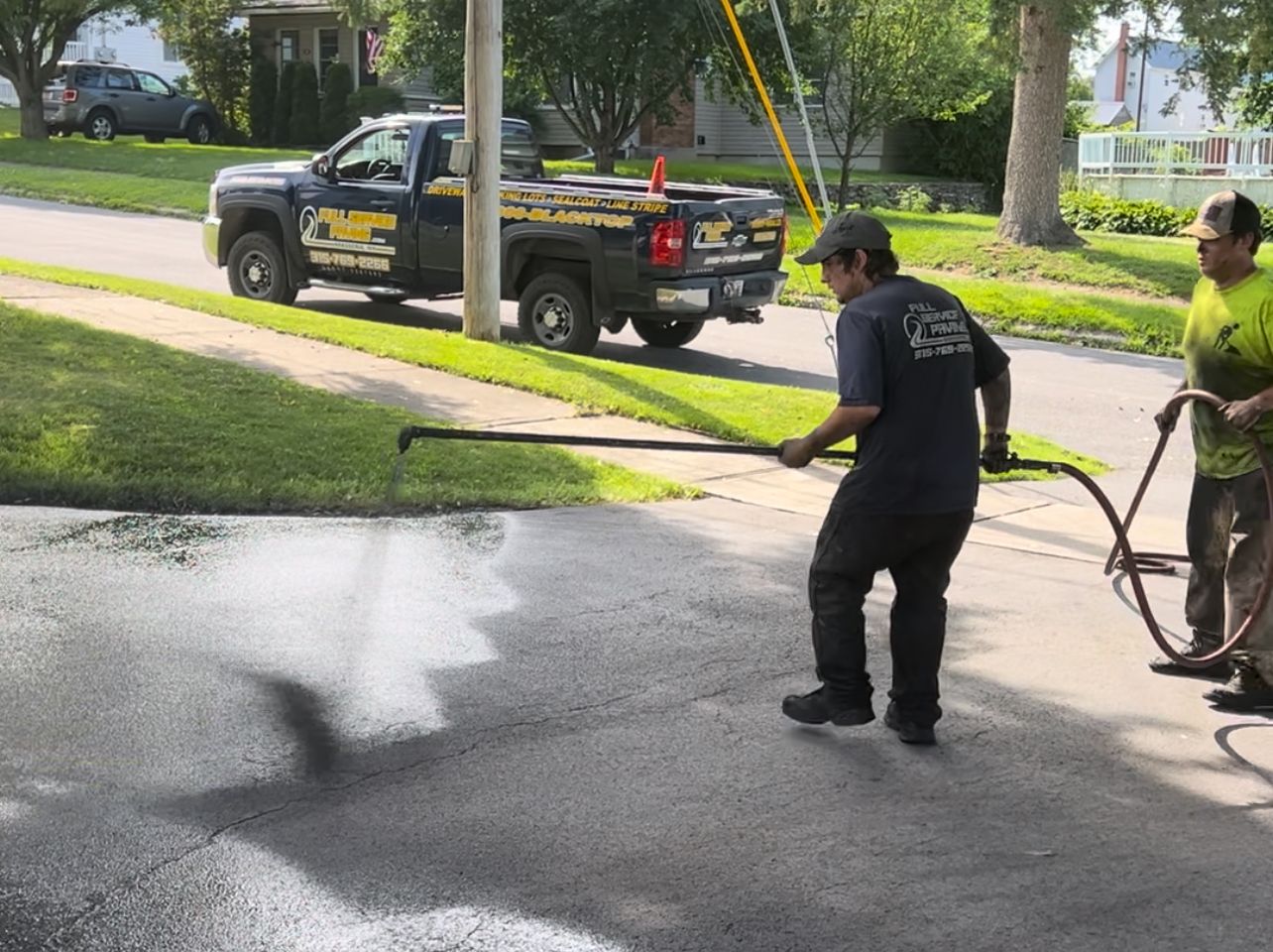 Two men are spraying asphalt on a driveway in front of a truck.