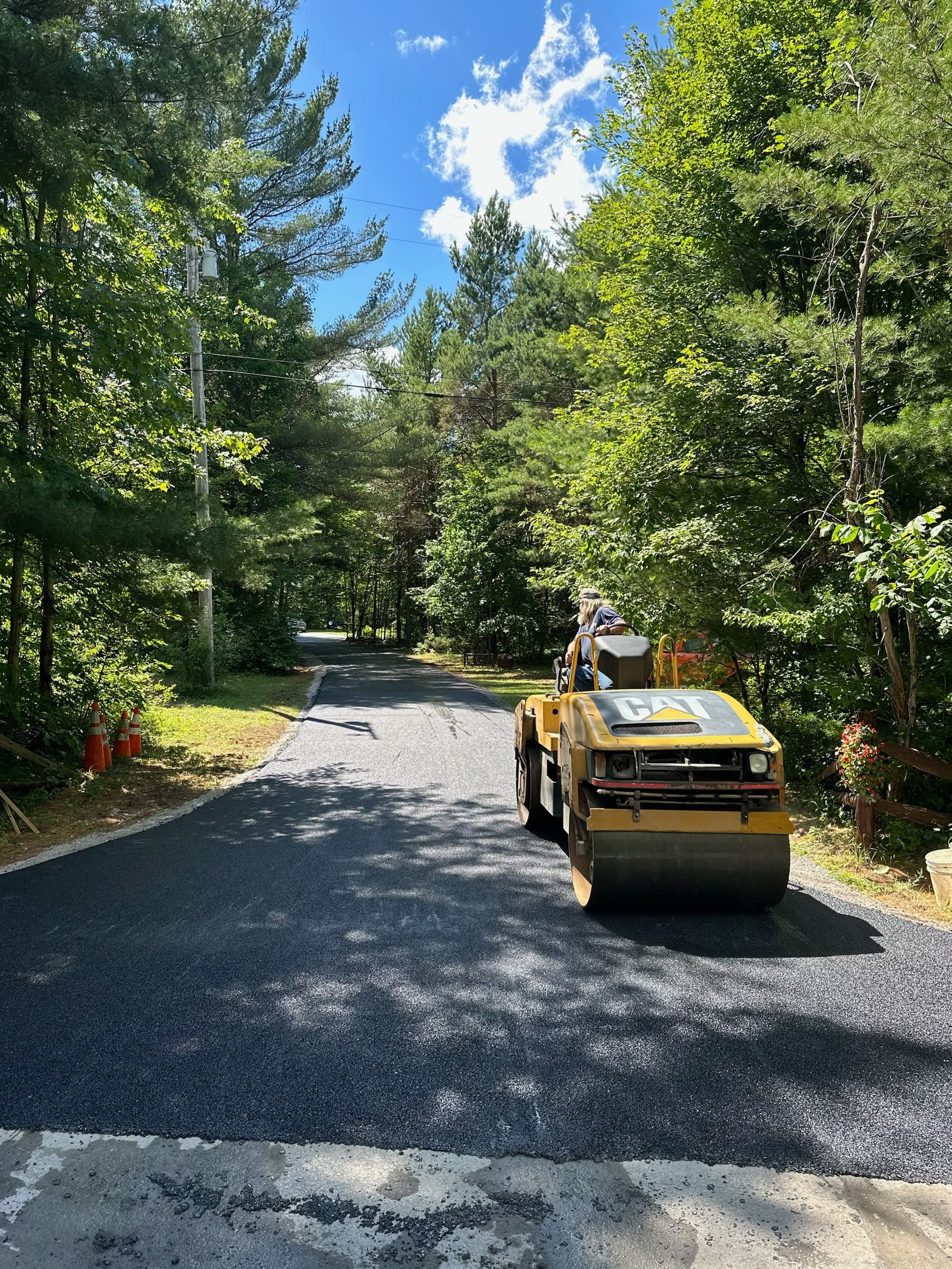 A yellow road roller is driving down a road surrounded by trees.