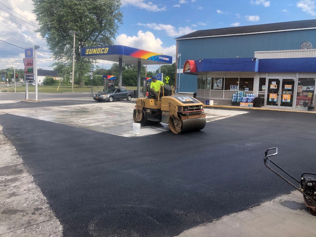 A man is driving a roller on a road in front of a gas station.
