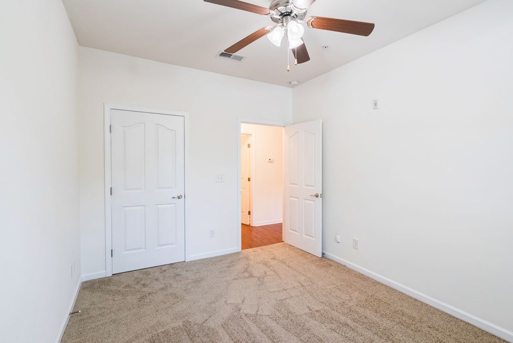 Empty bedroom with beige carpet and white walls, featuring a ceiling fan and two doors.