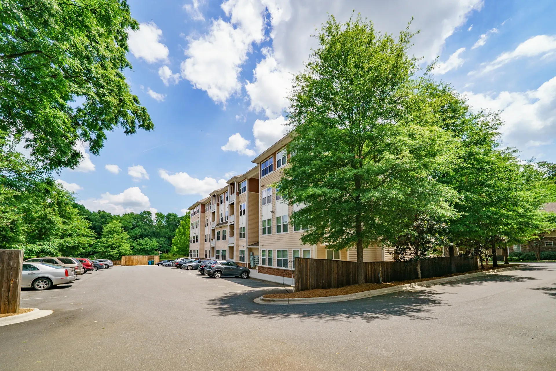 Exterior view of the apartment building alongside a parking area and trees under a blue sky with clouds.