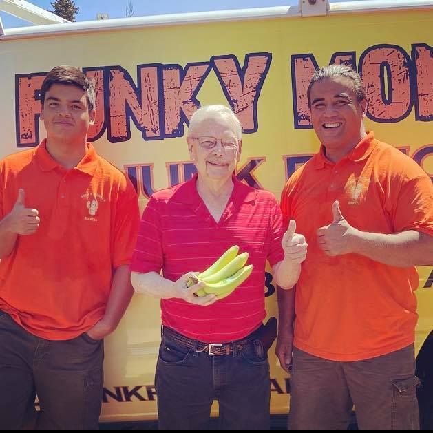 Three people in orange shirts with thumbs up, one holding bananas, in front of a yellow truck with 