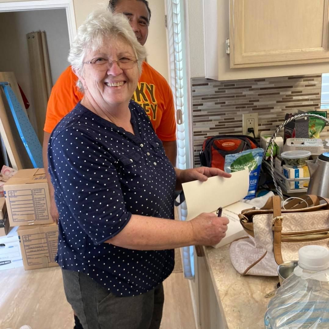 Woman signing document in kitchen, smiling, with man behind her. Boxes visible.