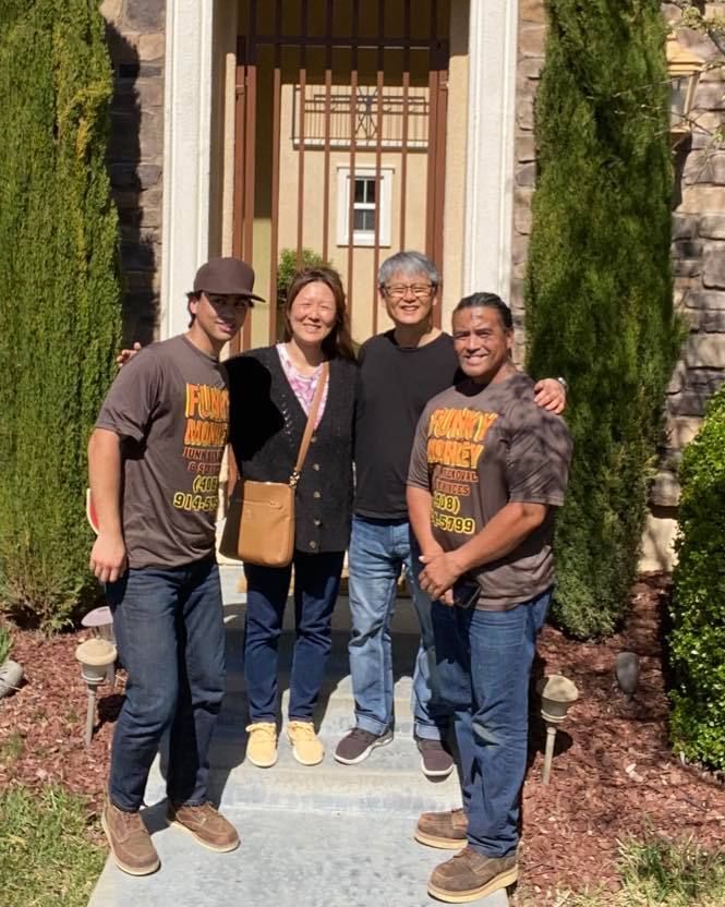 Four people standing in front of a house, smiling. Two men in brown shirts, jeans. A woman and another man in casual clothes.