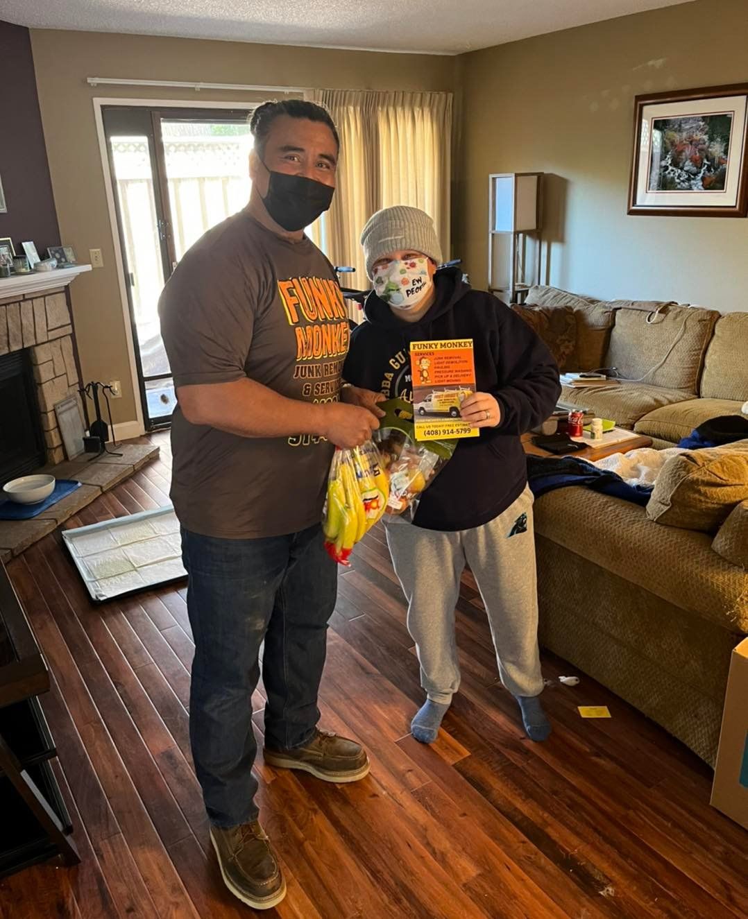 Man in mask handing gifts to a person wearing a mask and holding a book, inside a living room.