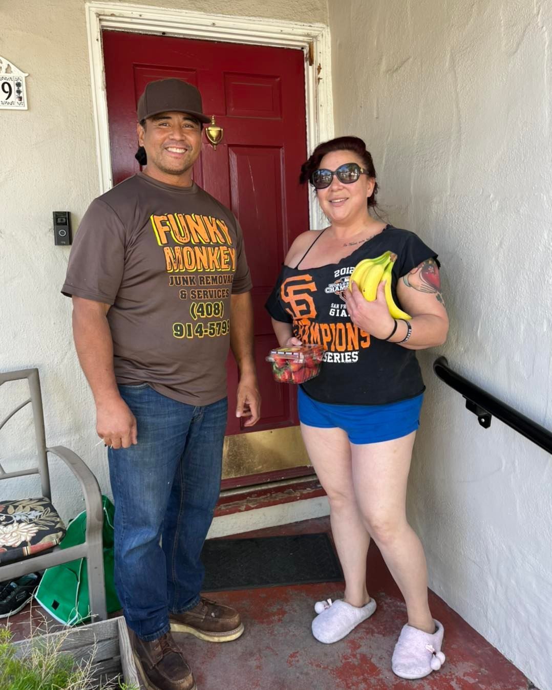 Two people stand in front of a red door. One holds bananas. The other wears a baseball team shirt.