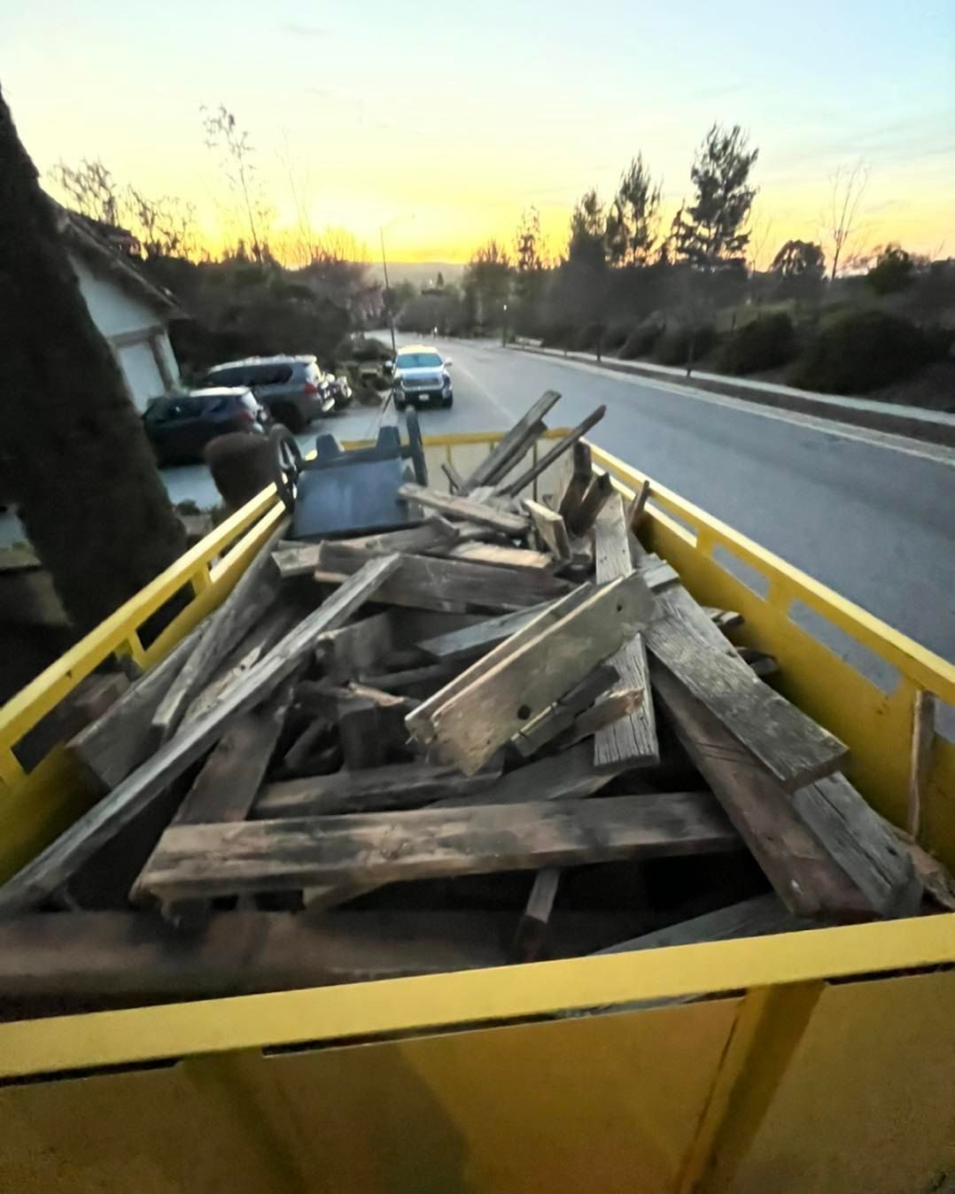 Yellow dumpster filled with wood debris on a residential street at sunset.
