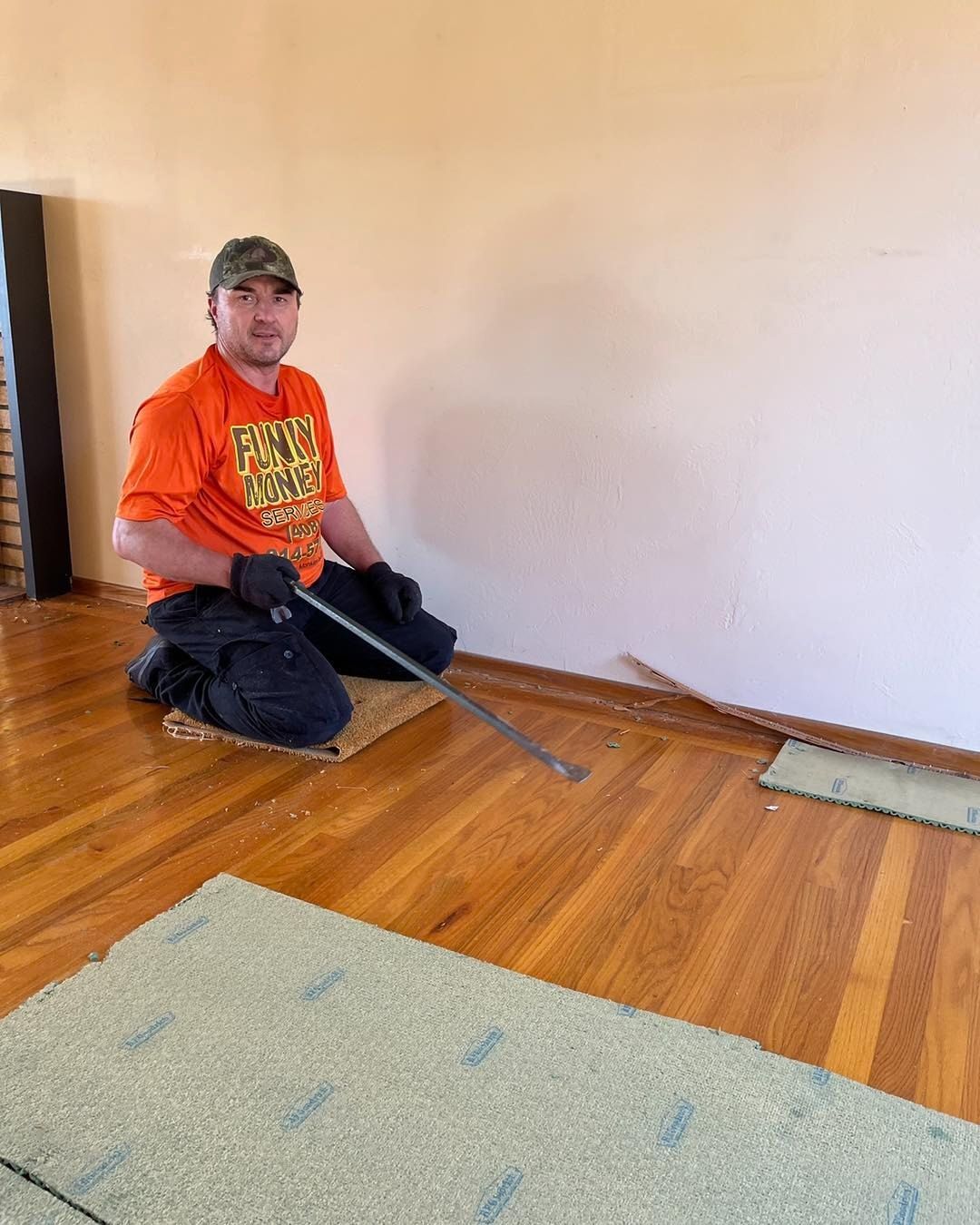 Man kneeling on wooden floor, removing trim with a pry bar. He wears orange shirt, cap.