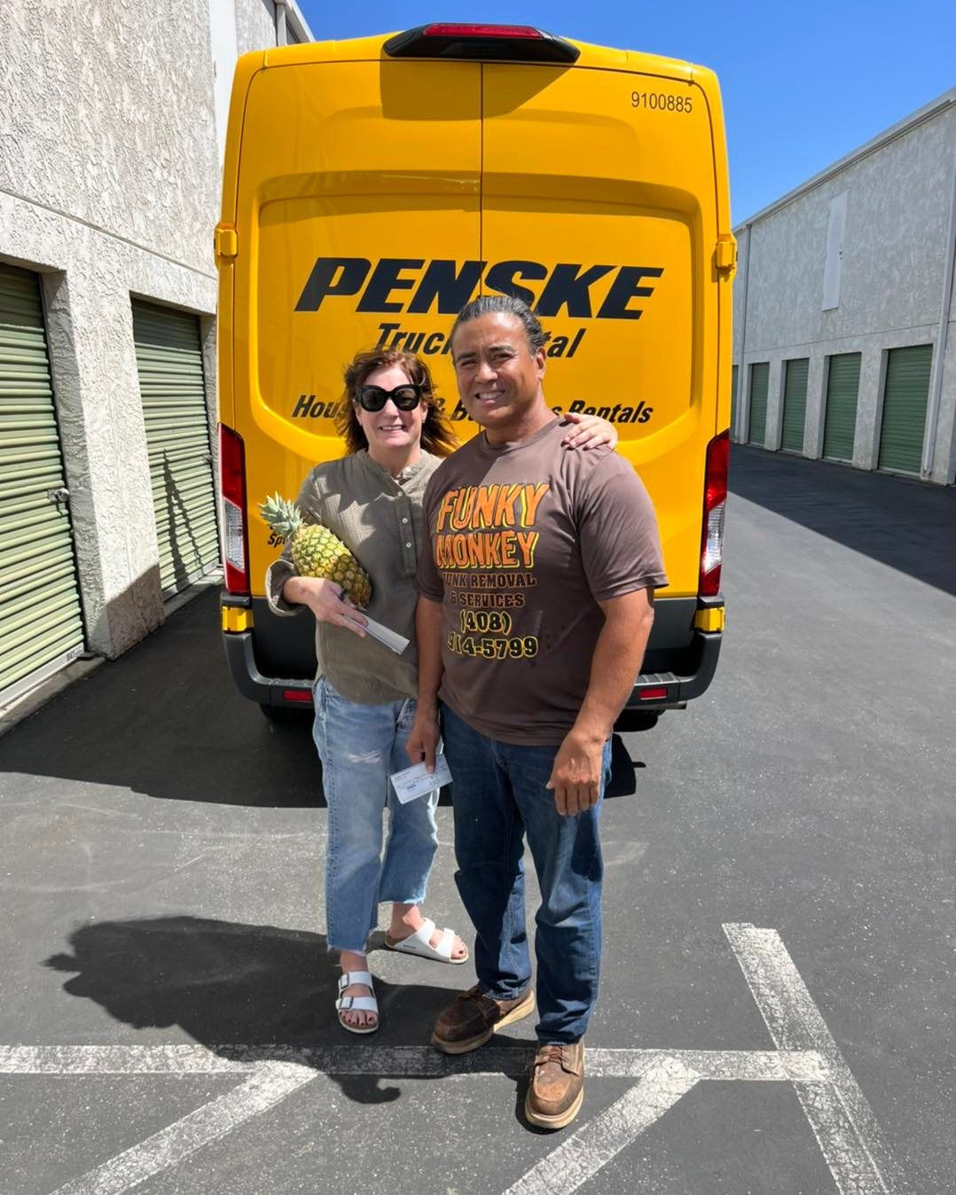 Two people stand in front of a yellow Penske truck. One holds a pineapple. Exterior setting.