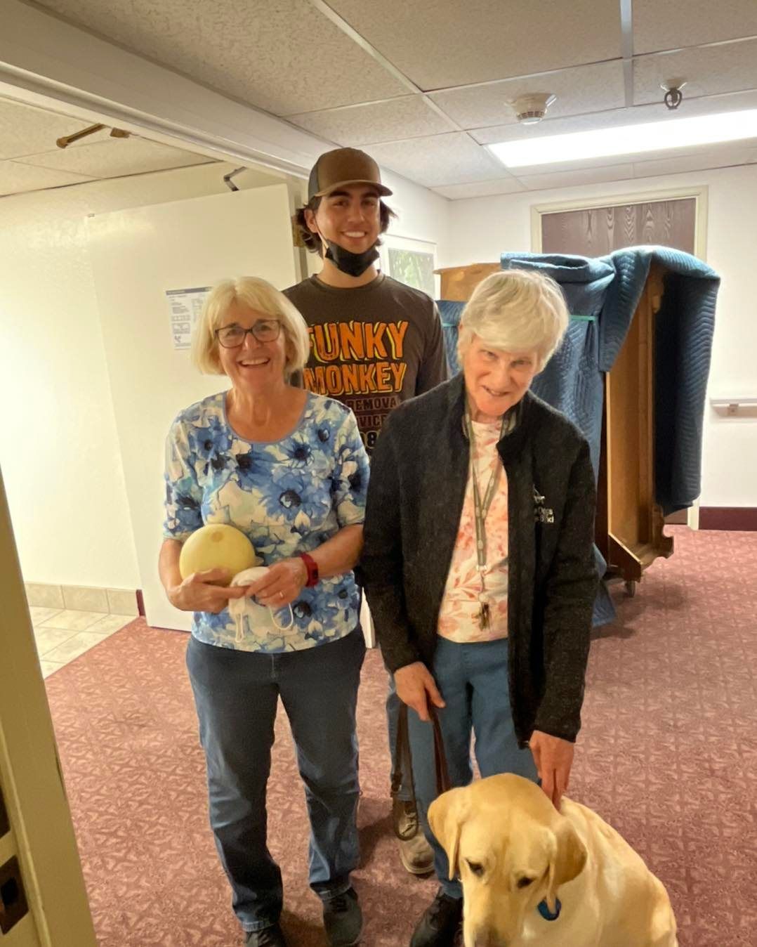 Two women and a man pose with a yellow lab inside a building. The women hold an item. The man wears a 
