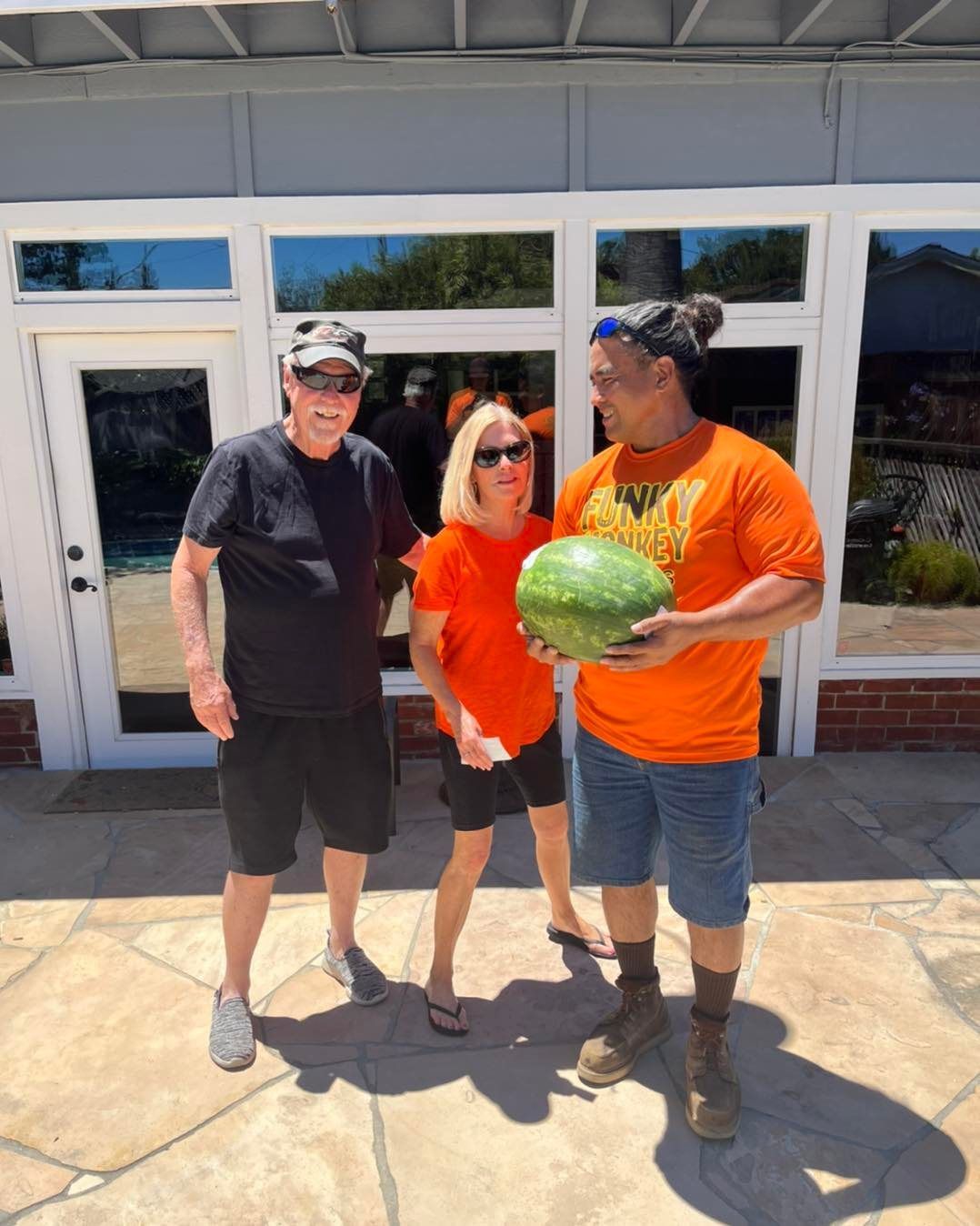 Three people pose outdoors; one holds a watermelon. They're in front of a building with large windows.