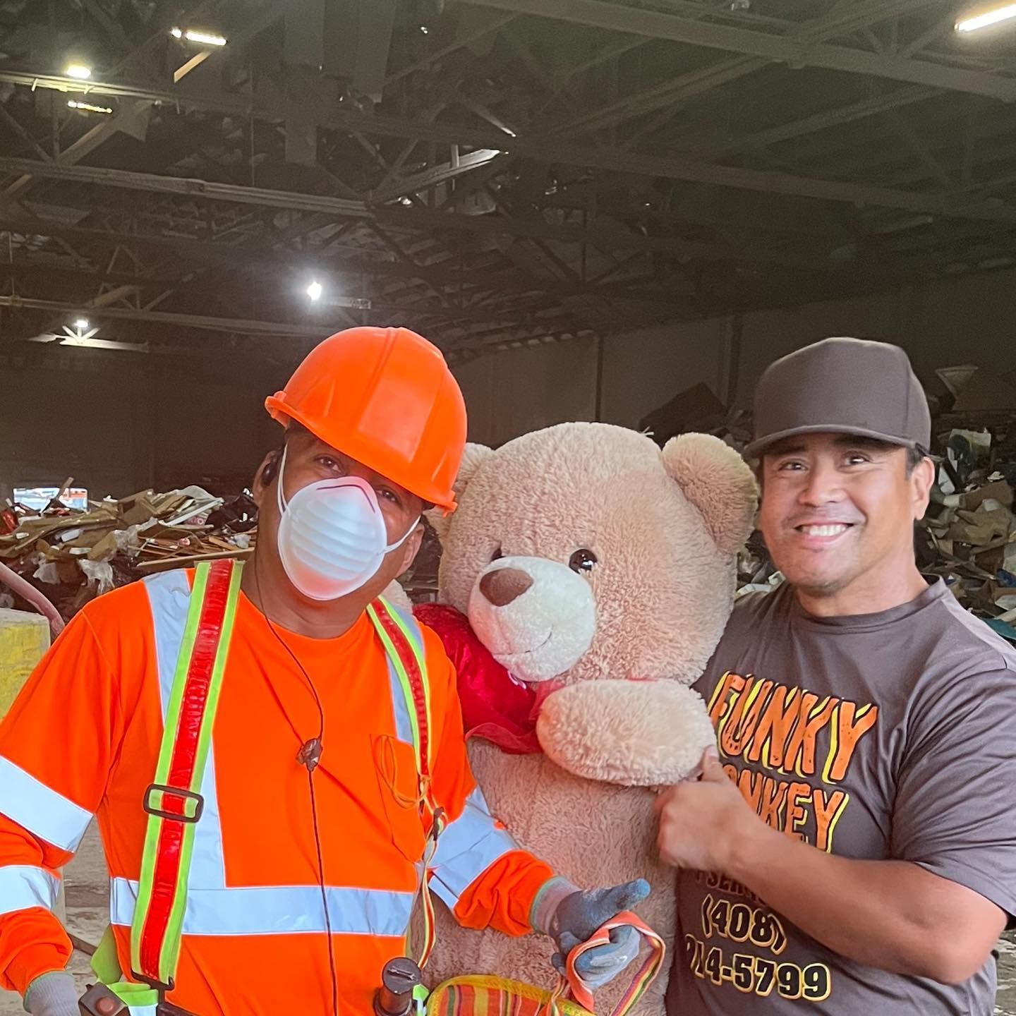 Two men pose with a large teddy bear in a recycling facility. One wears work gear; the other a baseball cap.