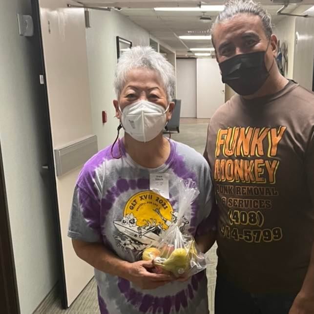 Woman in tie-dye shirt and man with mask pose. Woman holds fruit gift. Inside a building hallway.