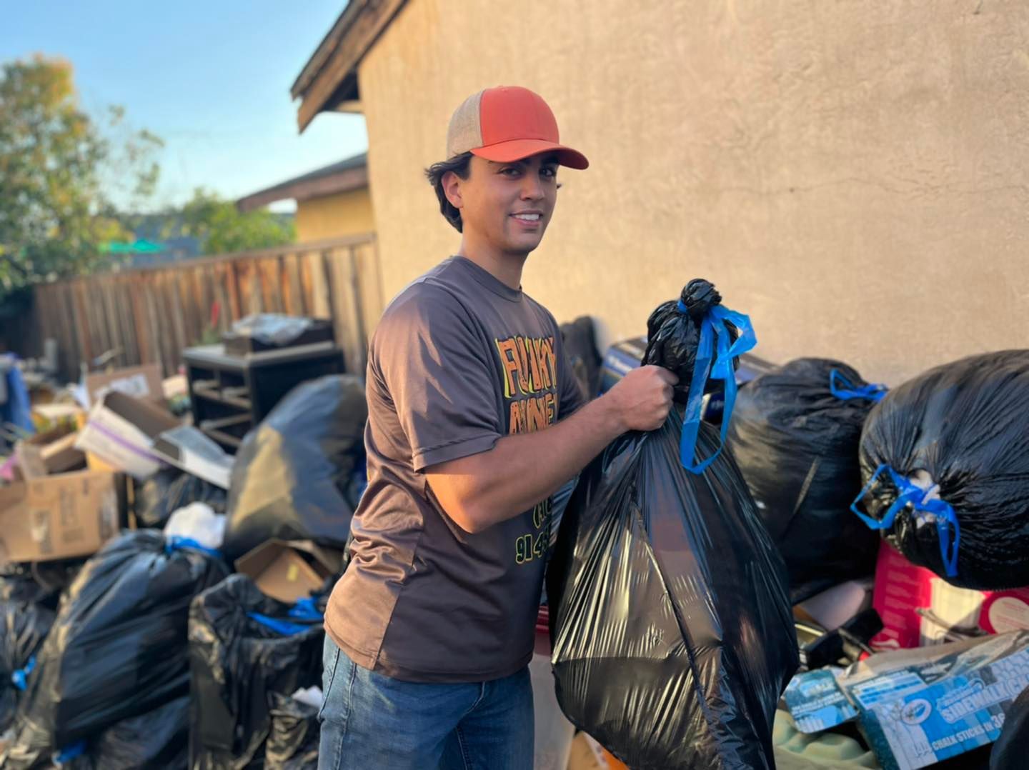 Man holding a black trash bag smiles near a pile of garbage bags. Brown shirt, red hat, sunny outdoor setting.