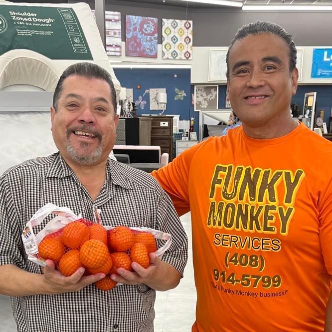 Two men smiling, one holding oranges in a mesh bag.  The other wears an orange shirt with 