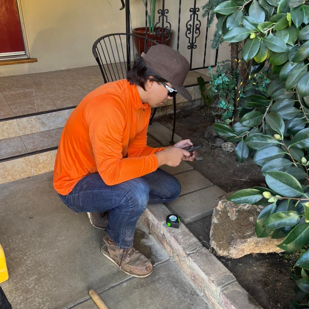 Man in orange shirt and hat crouches to measure a concrete border with a tape measure outdoors.